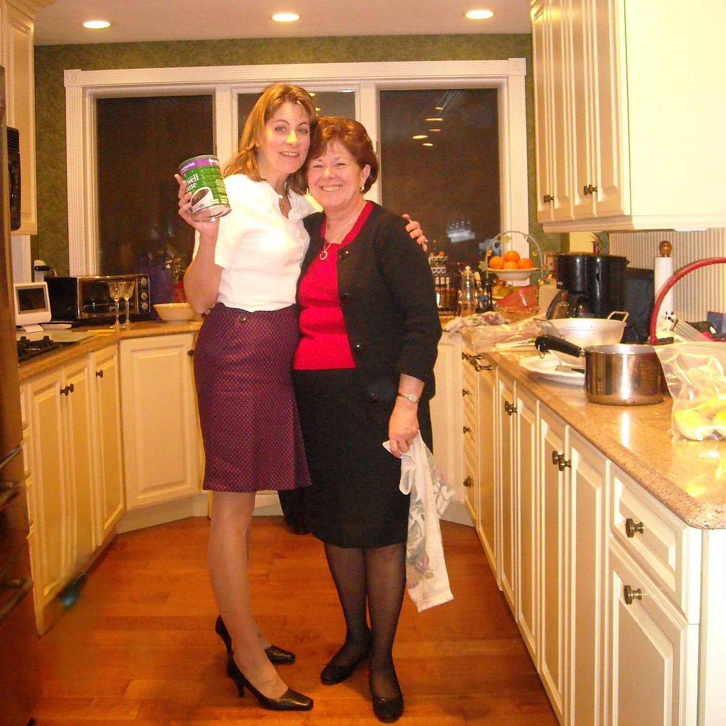 Two women, dressed for a holiday celebration, pose in the kitchen where they are surrounded by the remnants of a holiday meal