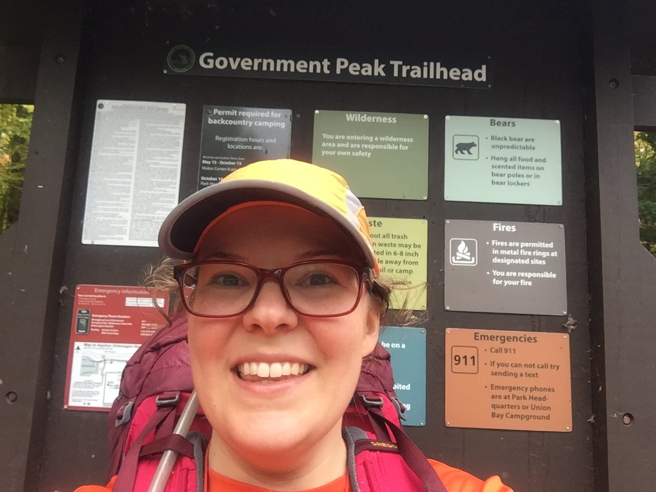 The author wearing an orange hat, red glasses, and a red backpack standing in front of the Government Peak Trailhead sign.