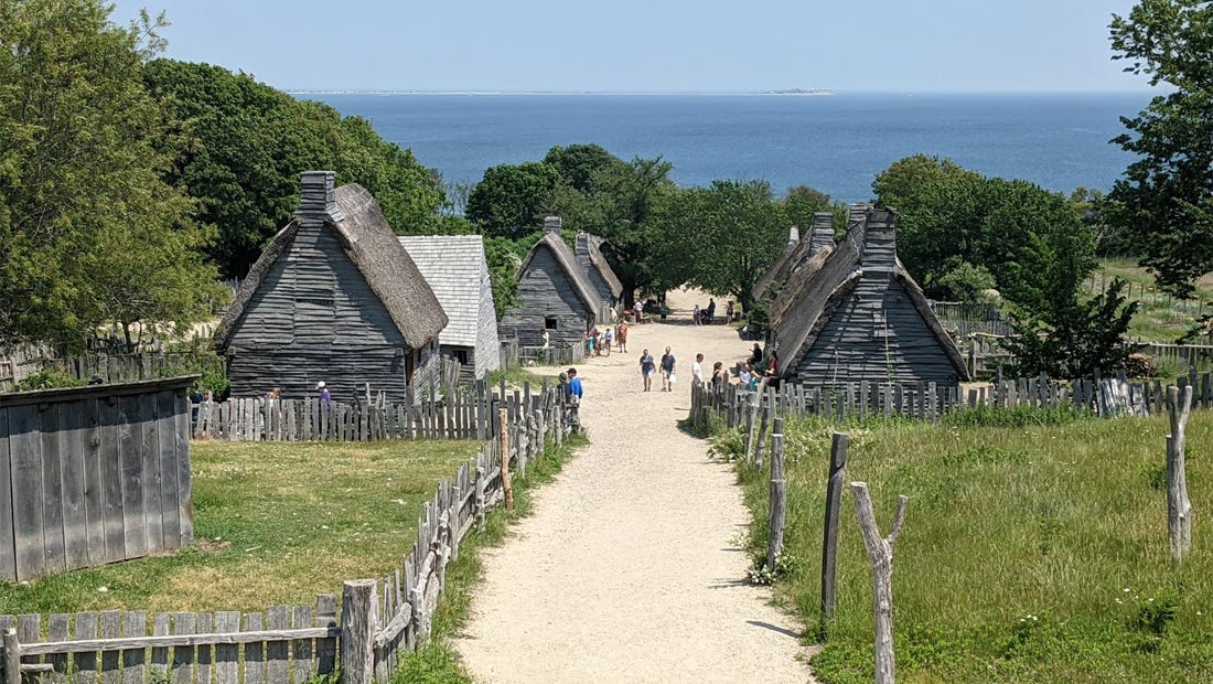 View down a dirt road of the roughly hewn homes of Plimoth settlers, the Atlantic Ocean in the not far distance.