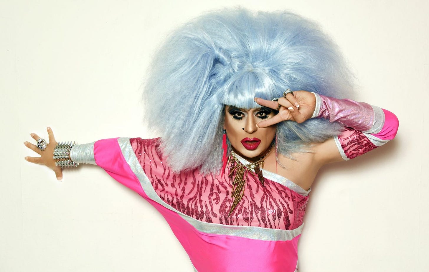 Against a white backdrop, a drag queen wearing a big icey blue wig and dramatic makeup poses with her arms, wearing a pink asymmetrical top. 
