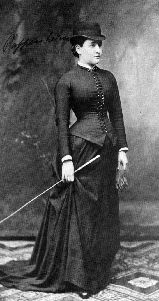 A black and white image of a white woman standing and looking off to the right in a hat, cinched waistcoat and long skirt.
