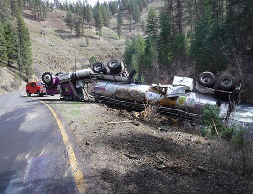 Photo of an overturned tractor-trailer rig at the side of a narrow mountain road, consisting of a purple truck, its passenger side roof almost flattened, and a tanker trailer that's badly dented from the crash.