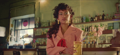 A woman with long curly hair in a pink suit slides a drink across the bar to the viewer