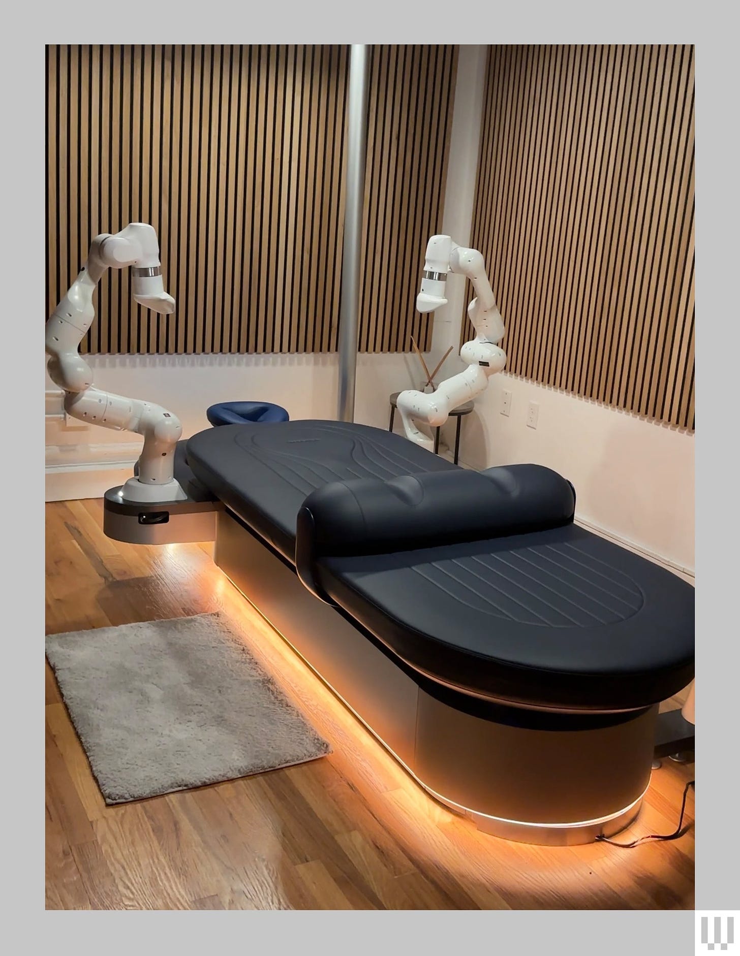 Black padded massage table in the center of the room with two angular white robotic arms hovering over the left and...