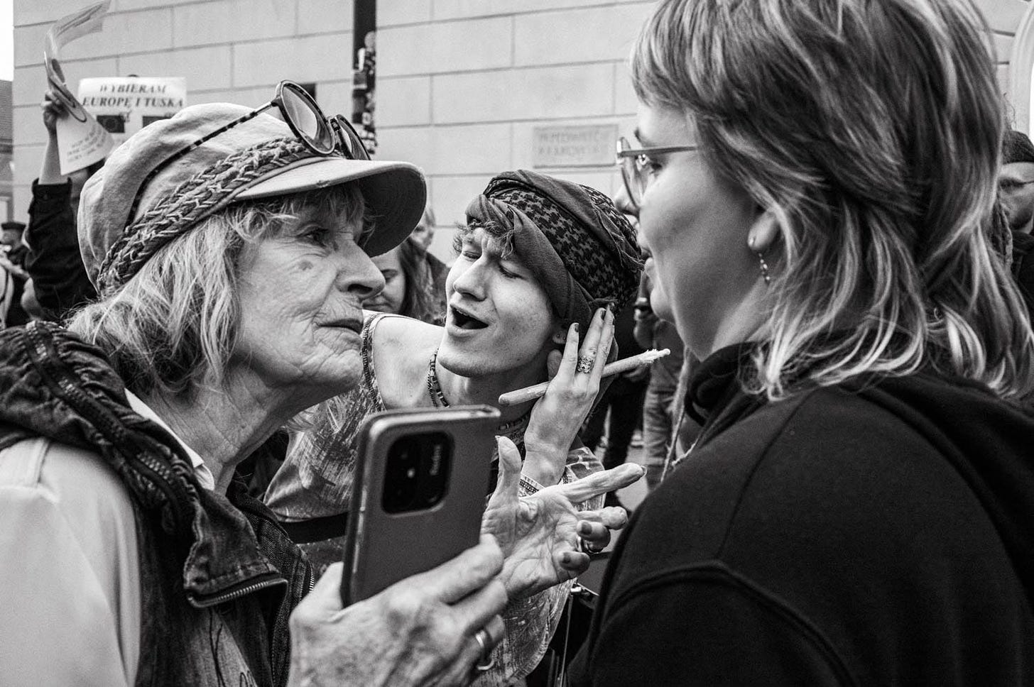 Three women at a protest or event. An elderly woman in a hat looks intently at a younger woman who is gesturing expressively. A third woman observes while holding a smartphone.