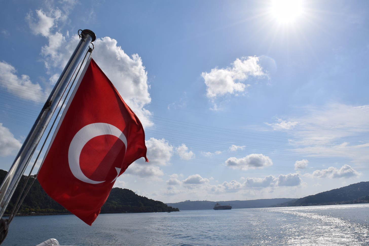 A Turkish flag hangs off a boat in the middle of a wide channel of water, beneath a big blue sky with a few clouds and bright sun.