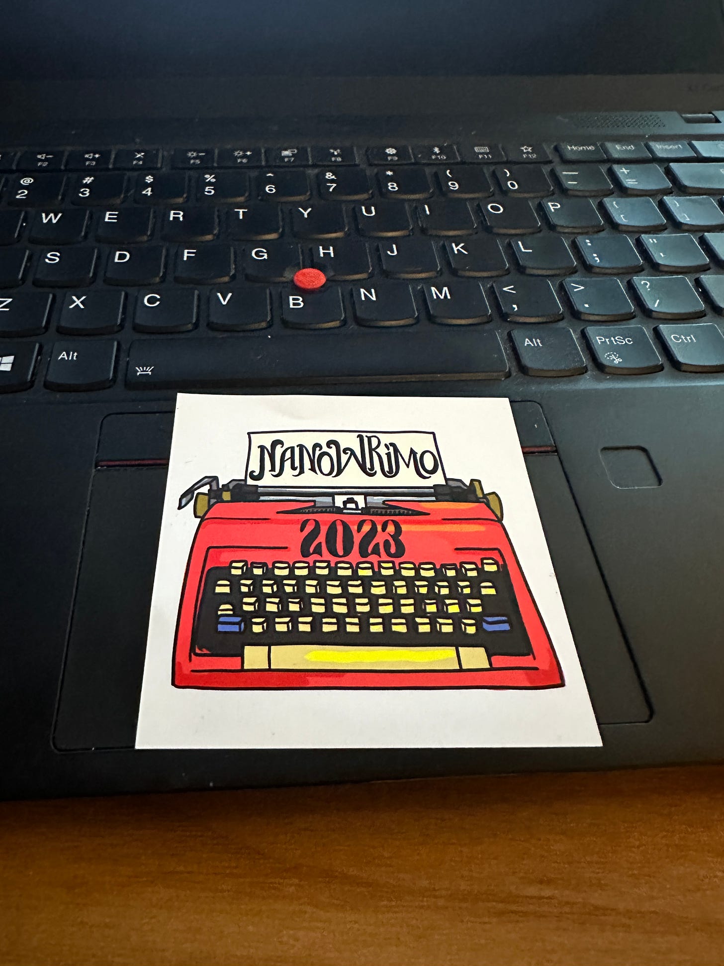 A Nanowrimo 2023 sticker showing a typewriter, resting on top of a laptop keyboard