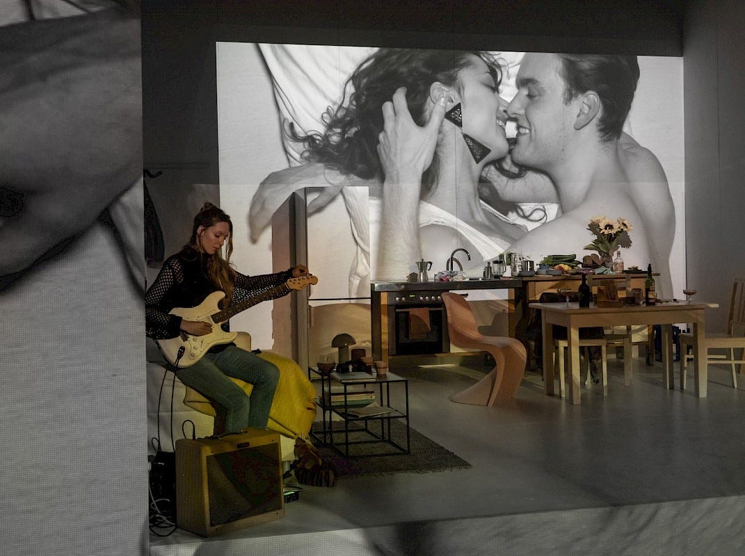 A woman plays a guitar in front of a screen in which a black and white image of a couple kissing can be seen.