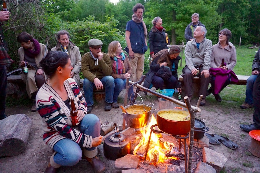 Group of people around a campfire. Some sitting on low wooden benches, others standing nearby.