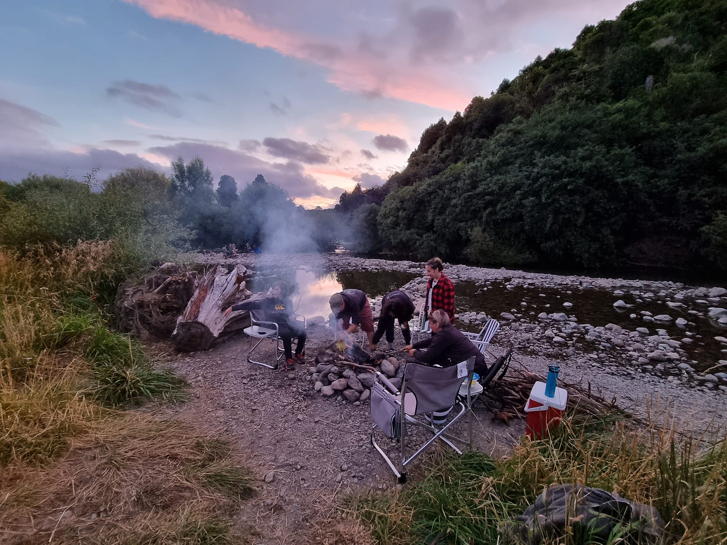 A few people making a fire on a riverbank in the sunset.