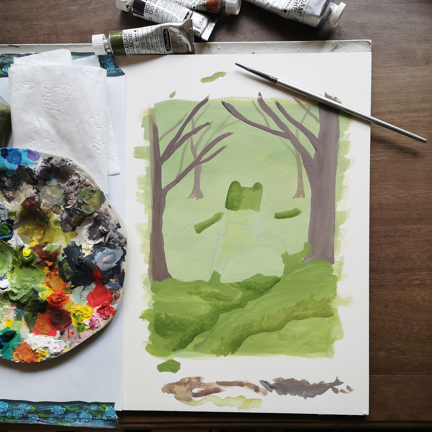 A paint palette and some tubes of gouache lie beside a painting in progress, which has a green background and what appears to be a jaunty green frog marching along the mossy ground beneath some trees.
