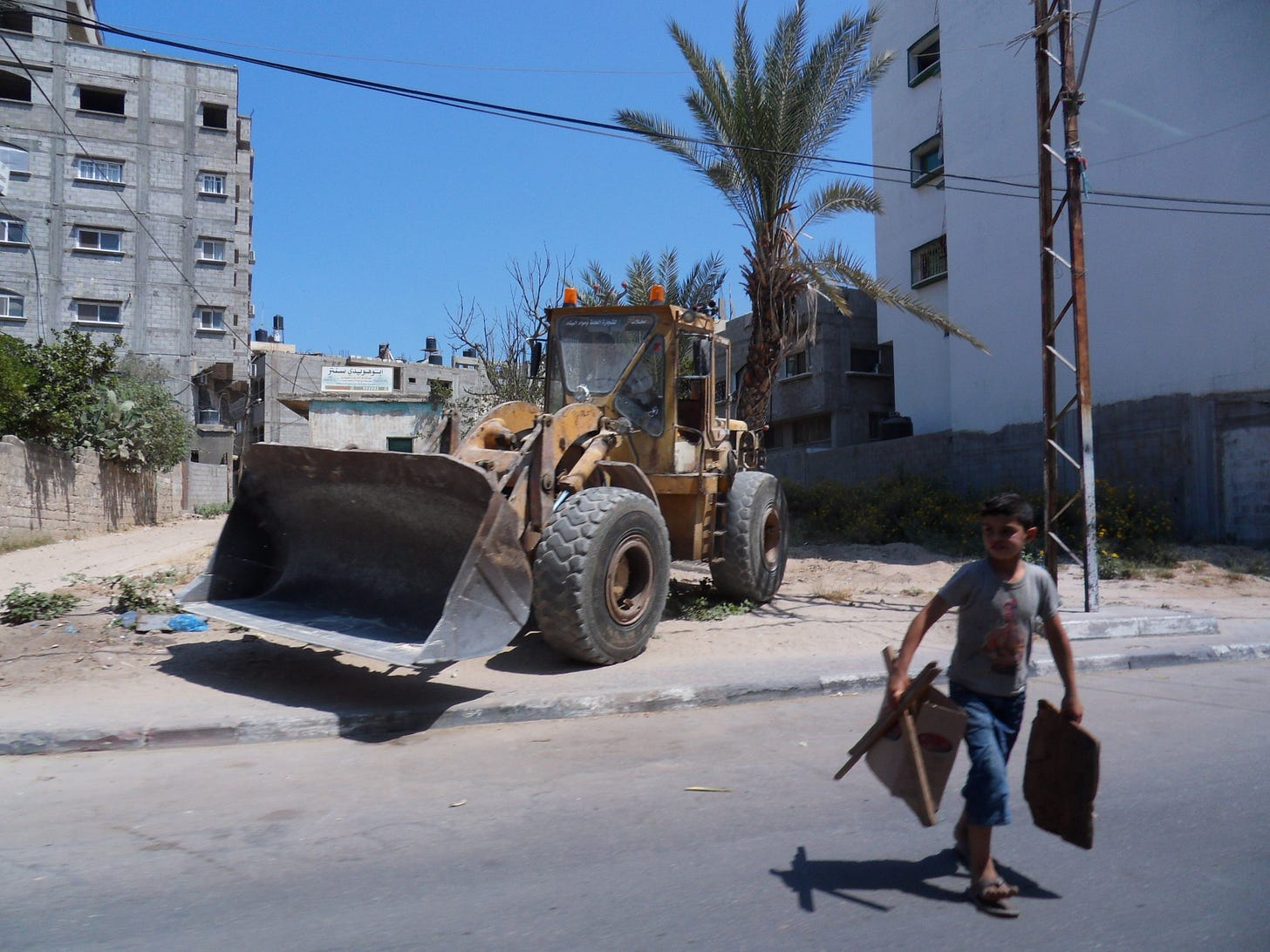 A boy carries wood and cardboard scraps across a road, and behind him sits a large bulldozer