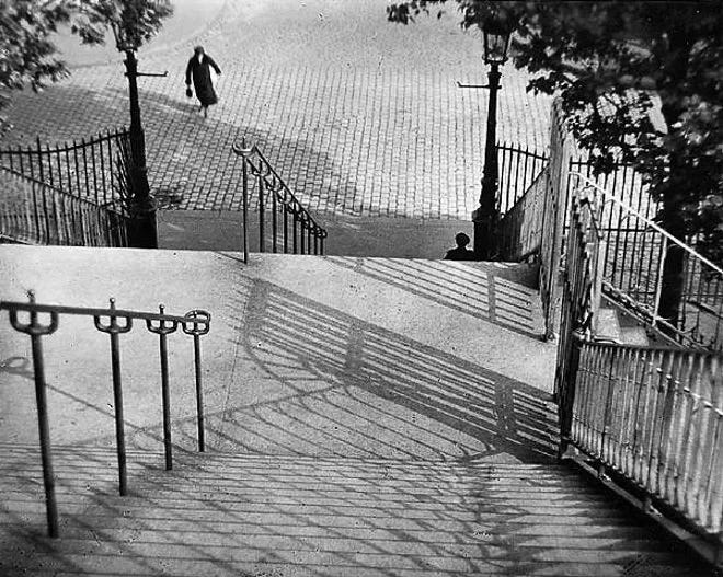 a photo by André Kertész copyright his state. It shows figures on a street from a high viewing angle looking down some stairs with railings. It features shape and form, shadows and angles.