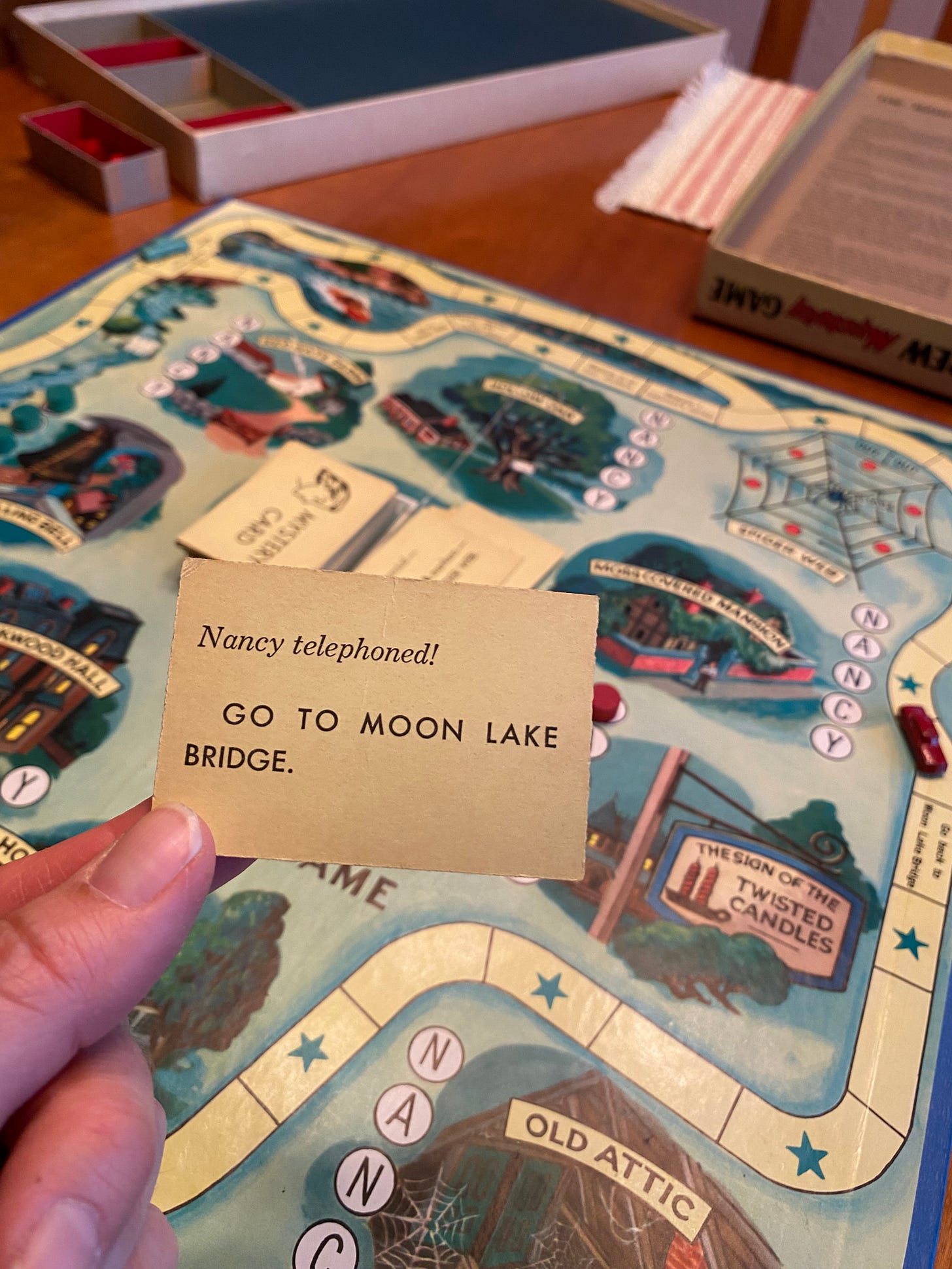 Image of the Nancy Drew Mystery Game from the 1940s, hand holding a card that says "Nancy telephoned! Go to Moon Lake Bridge."