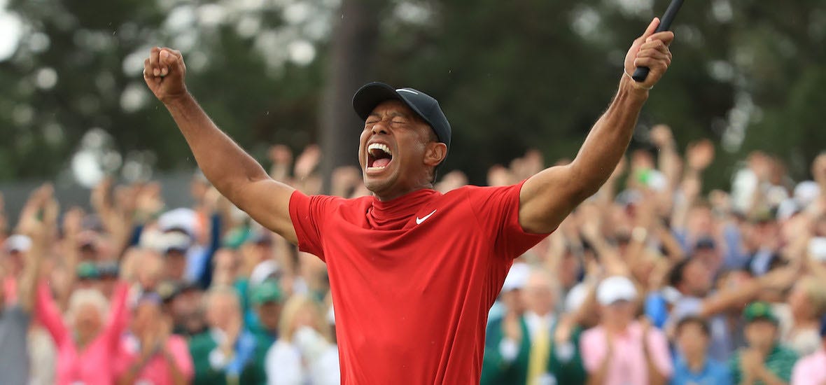 Tiger Woods celebrating with arms raised in victory, wearing his iconic red Nike shirt and black cap, with cheering crowd in the background.