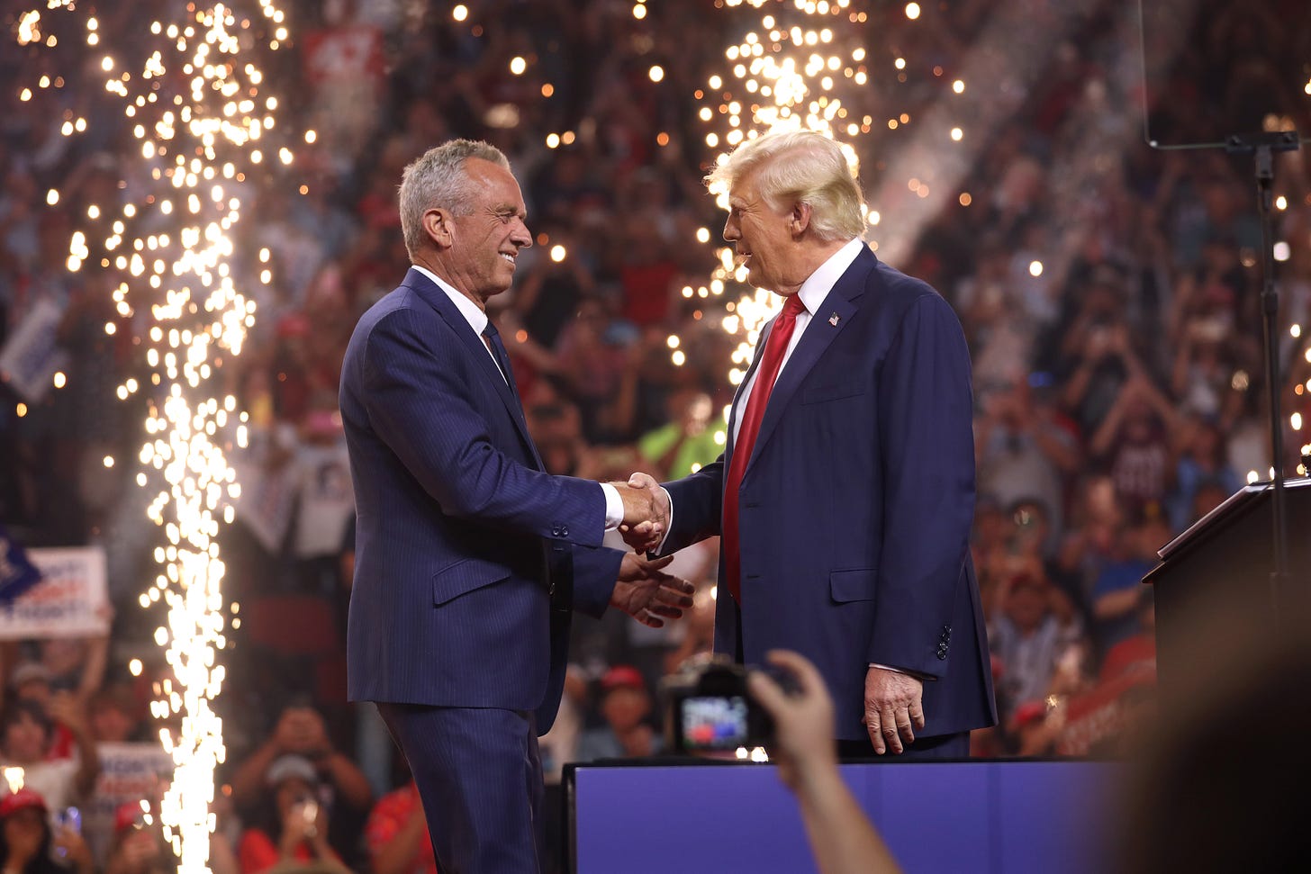 RFK Jr. and Trump shake hands on a stage with fireworks behind them at a political rally. A crowd and cameras are visible in the background. RFK Jr. and Trump shake hands on a stage with fireworks behind them at a political rally. A crowd and cameras are visible in the background.