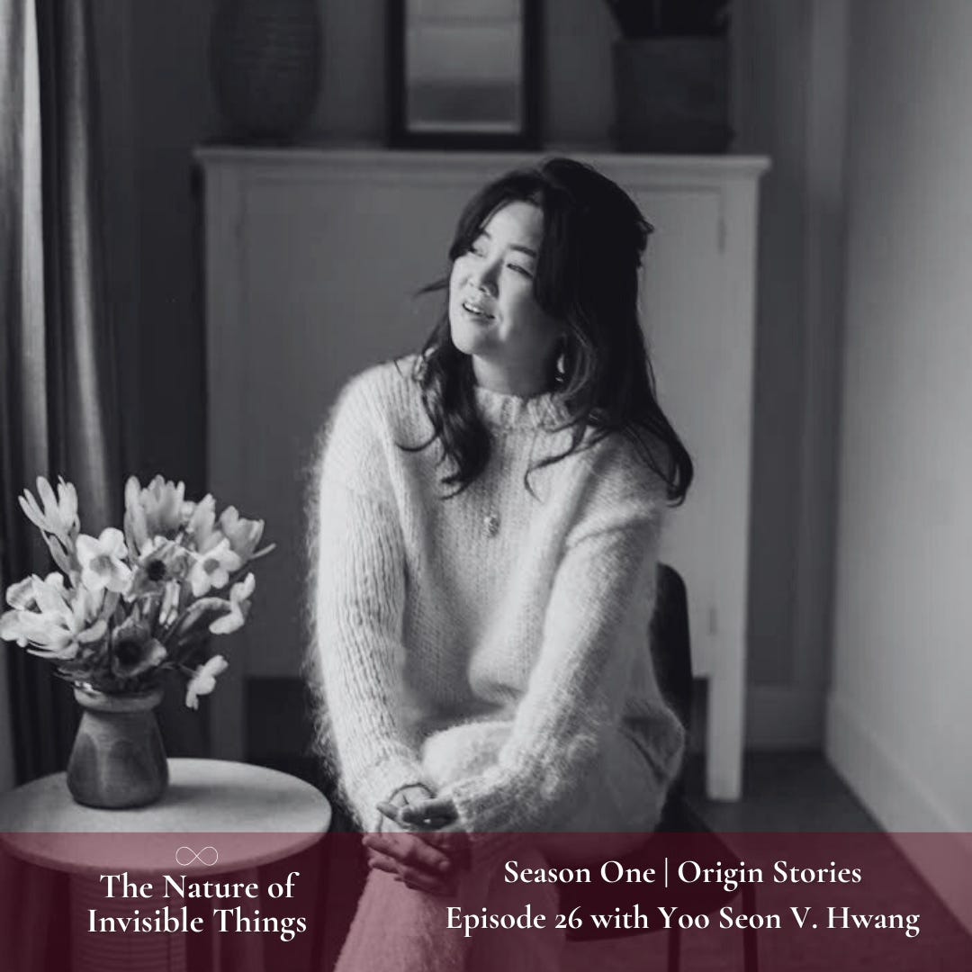 Yoo Seon, a Korean woman, is sitting in a chair with her legs crossed looking out a window. She has dark hair that is half-up with her bangs framing her face. She is wearing a light colored fuzzy sweater. To her side is a small side table with a vase full of flowers. She has a dreamy expression on her face.