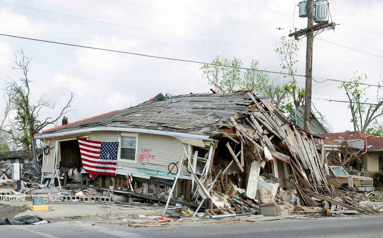 a long grey building badly damaged by hurricane. the front is torn off, with many planks of wood and debris collapsing forward into a pile. a large window has been blown away. many shingles are missing from the roof. the side of the house has been spray painted with red and black signs to indicate search-and-rescue status. an American flag has been attached backwards to the side of the house. cement blocks and debris litter the area around the house.