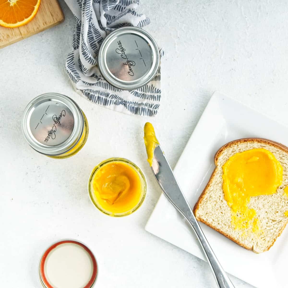 Small mason jars of orange curd next to a plate with slice of bread spread with the orange curd.