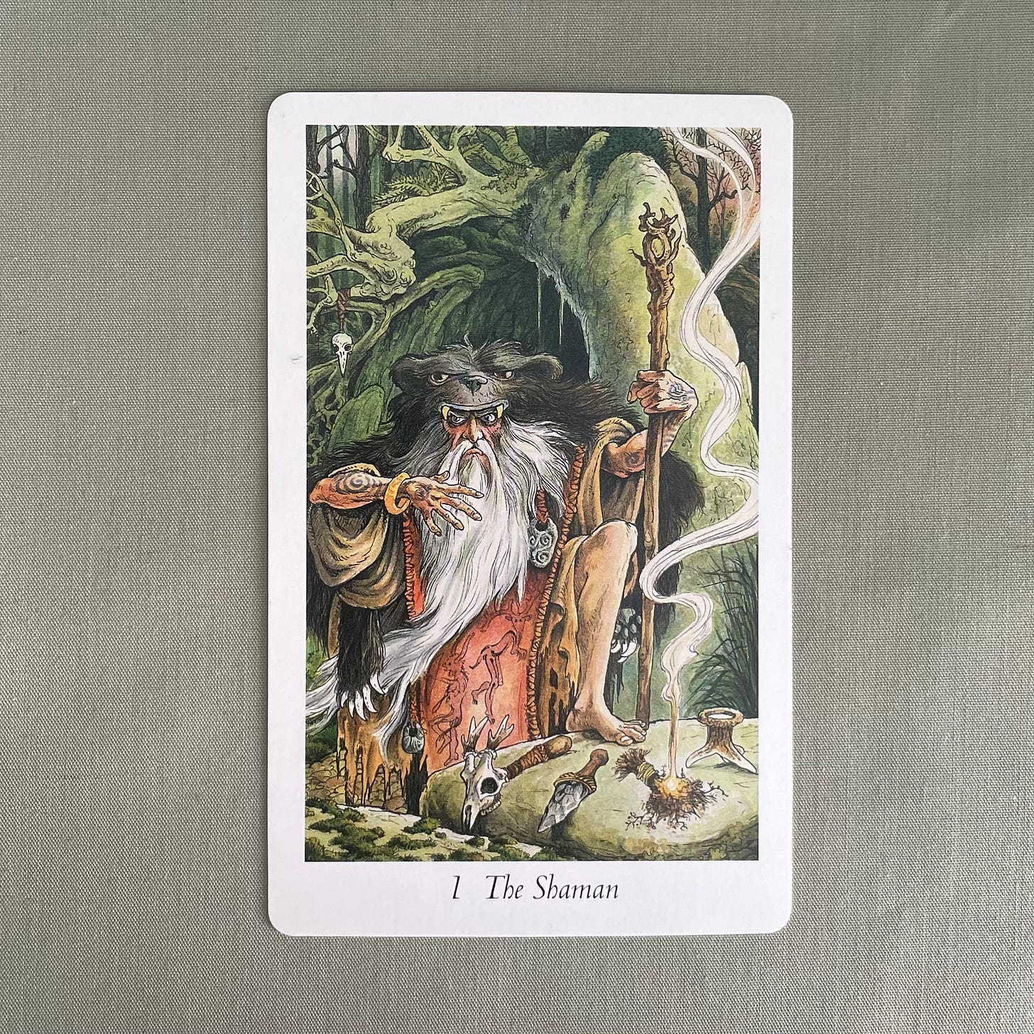A hand drawn tarot card sits on a sage green cloth. The card depicts a wildly bearded old man dressed in a bear cloak (the bear’s head forming the hood) and various other animal skins. His arms are tattooed with spirals and he wears a gold torc on his right wrist. In his left hand is a gnarled wooden staff and in front of him, on a stone table, are various items for performing magic with the elements, a roebuck skull rattle for air, a flint knife for earth, a lit bundle of herbs for fire and a hollow antler-tine cup for water. Behind him are the ancient twisted trees of the wild wood, and it appears to be either dawn or dusk from the reddish tint to the small amount of sky visible at the top right of the card.