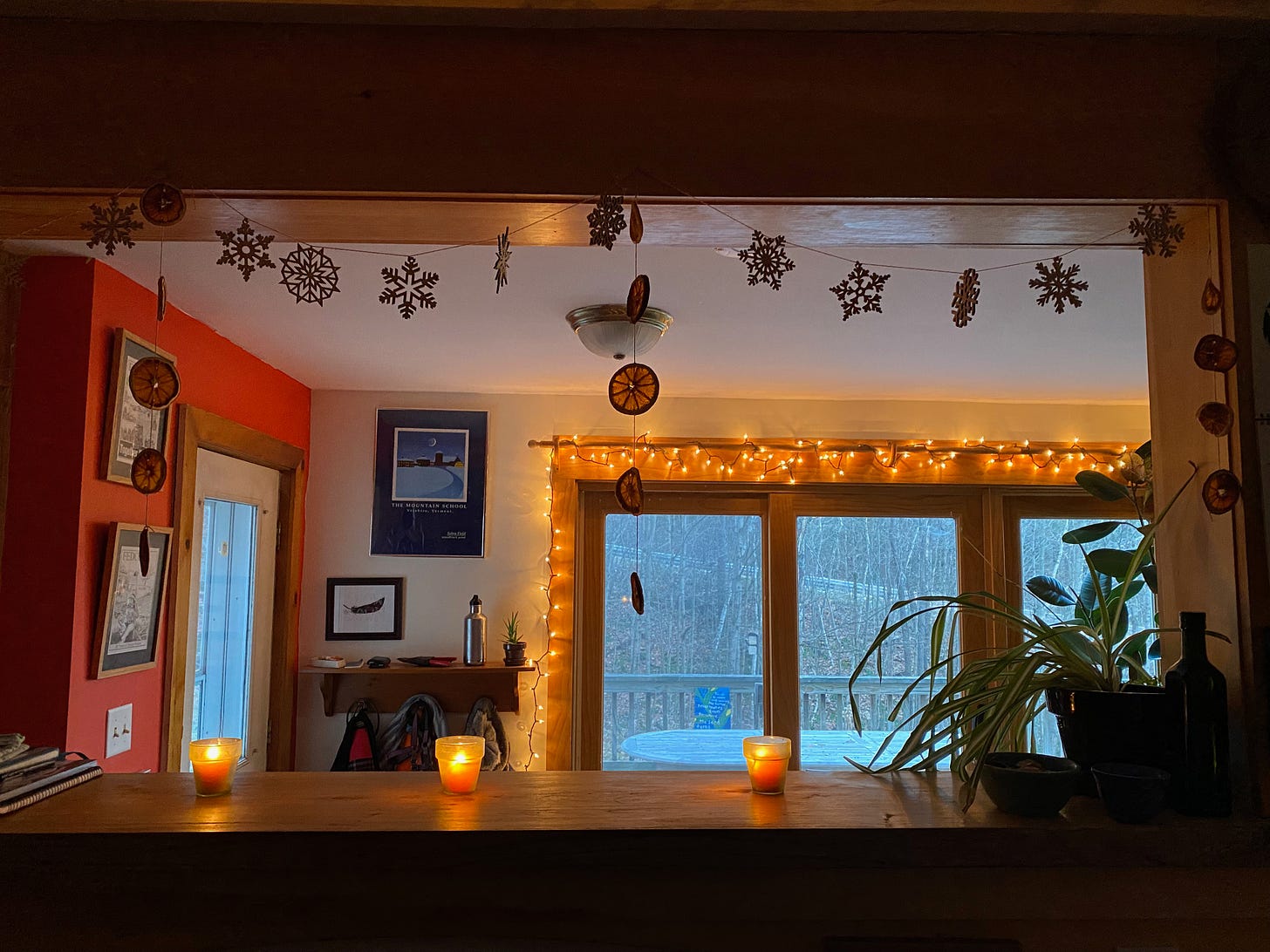 Wooden snowflakes and dried orange garlands hanging above a kitchen counter. There are lit candles on the counter and twinkling lights strung along the windows.