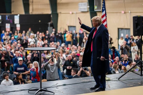 Donald J. Trump, wearing a long jacket, waves to a crowd from a stage.