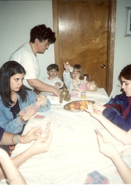 A photo from the 90s shows a family around a table after dinner. Two little girls - Cass and her sister - are at the end playing with dolls. There are others playing cards.