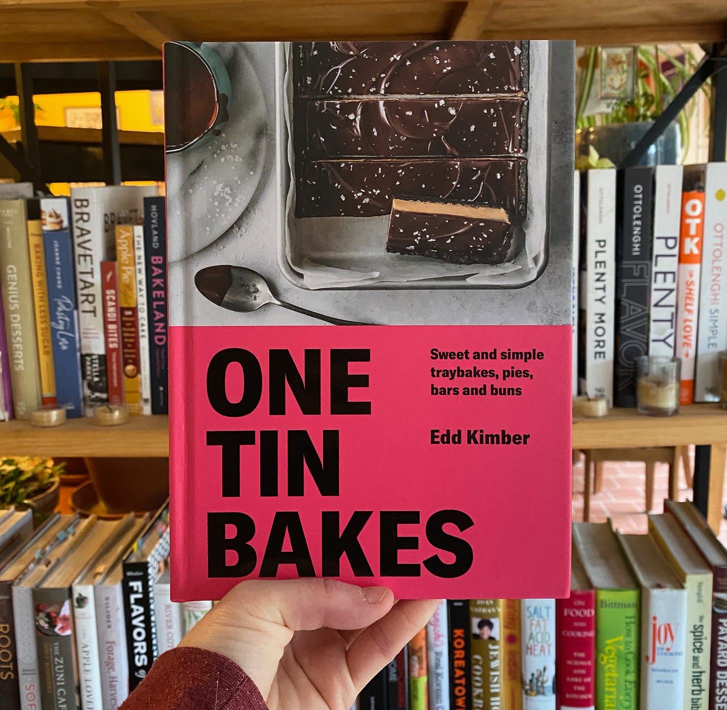 My hand holding One Tin Bakes in front of a shelf full of cookbooks.