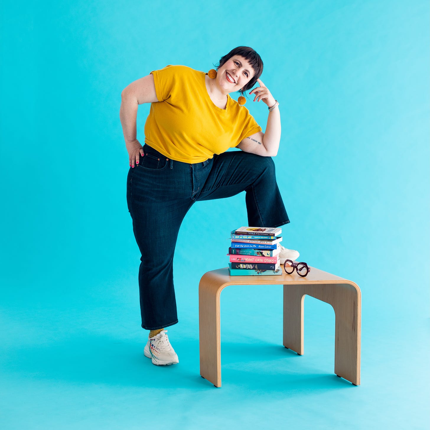 Lauren wearing a yellow shirt and jeans with her foot up on a table that has a pile of queer books on it and her big rimmed glasses