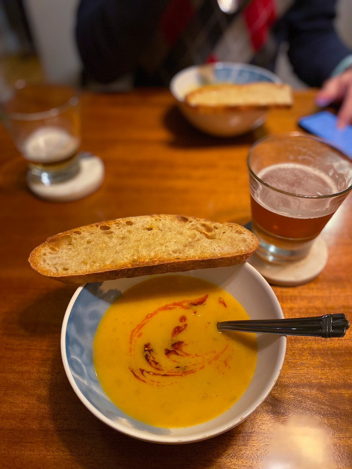 A bowl of the blended carrot soup described above, with a swirl of chili oil on top and a slice of bread at the edge of the bowl. A glass of beer is on the coaster to the right, and Jeff is across the table with his own bowl.
