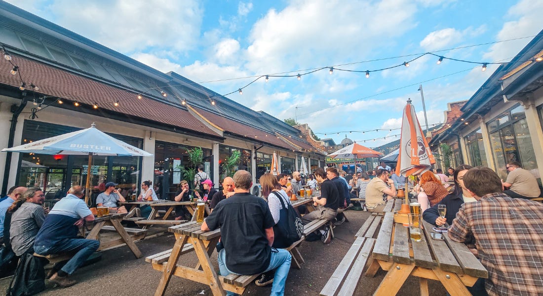 An outdoor dining spot with people sat on benches with umbrellas on, string lights over them, and two buildings either side