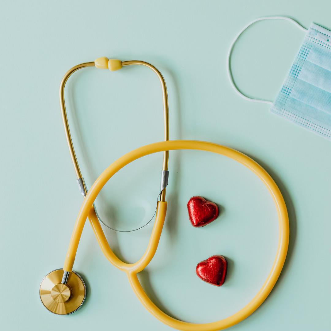 A photo of a stethoscope and medical mask laying on a very light blue background. The stethoscope is yellow and there are 2 red, heart shaped chocolates.