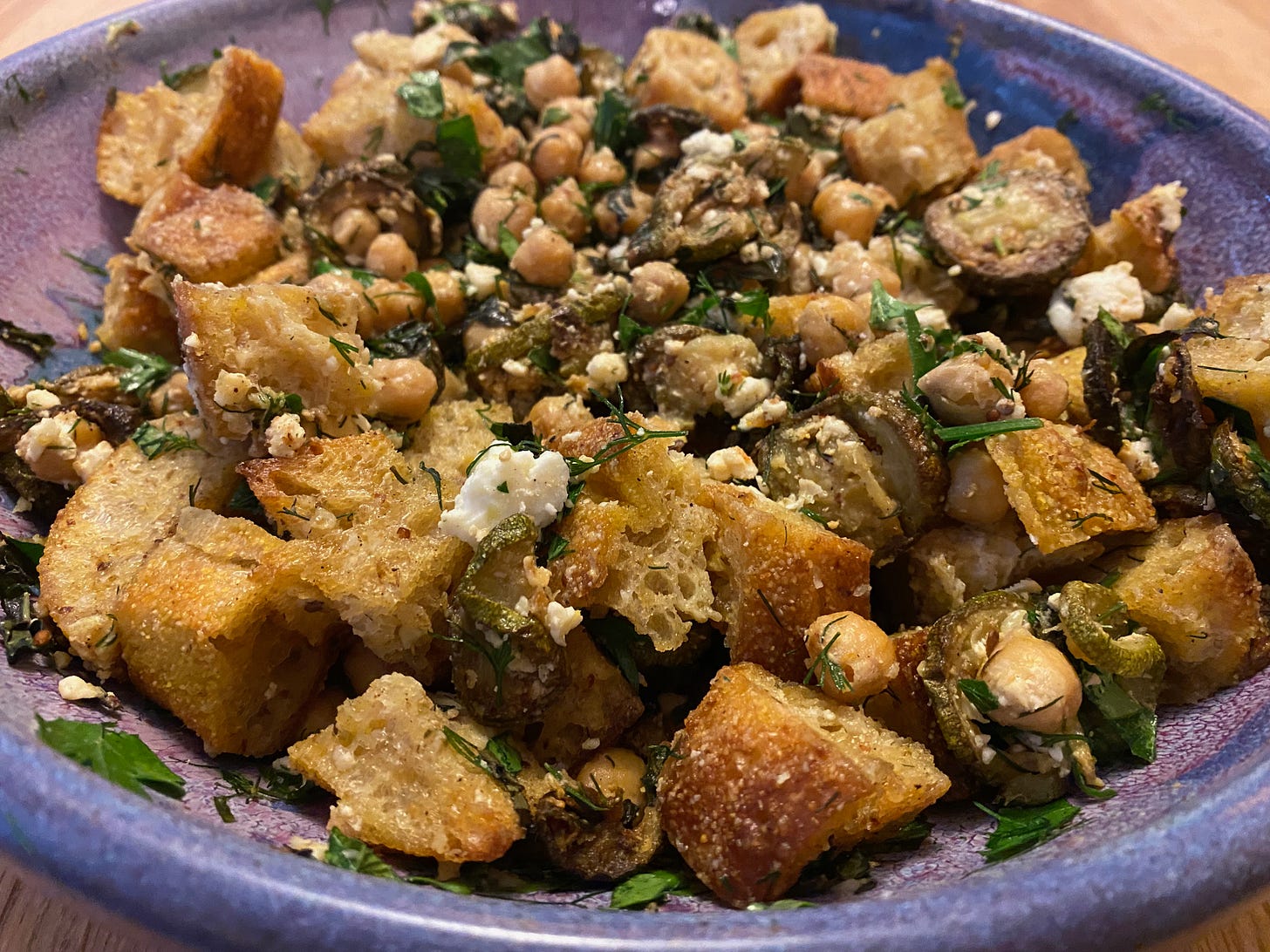 Closeup of a purple ceramic serving bowl filled with crispy fried bread, chickpeas, chopped herbs, and crumbled feta.