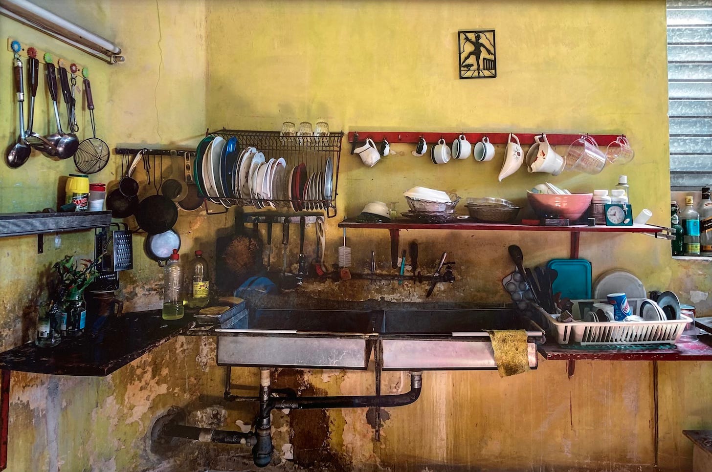 Photographic art made by Leslie Osterling of the interior of a Peruvian kitchen showing twin metal sinks and kitchen utensils hanging on the yellow walls and on the wooden countertops.