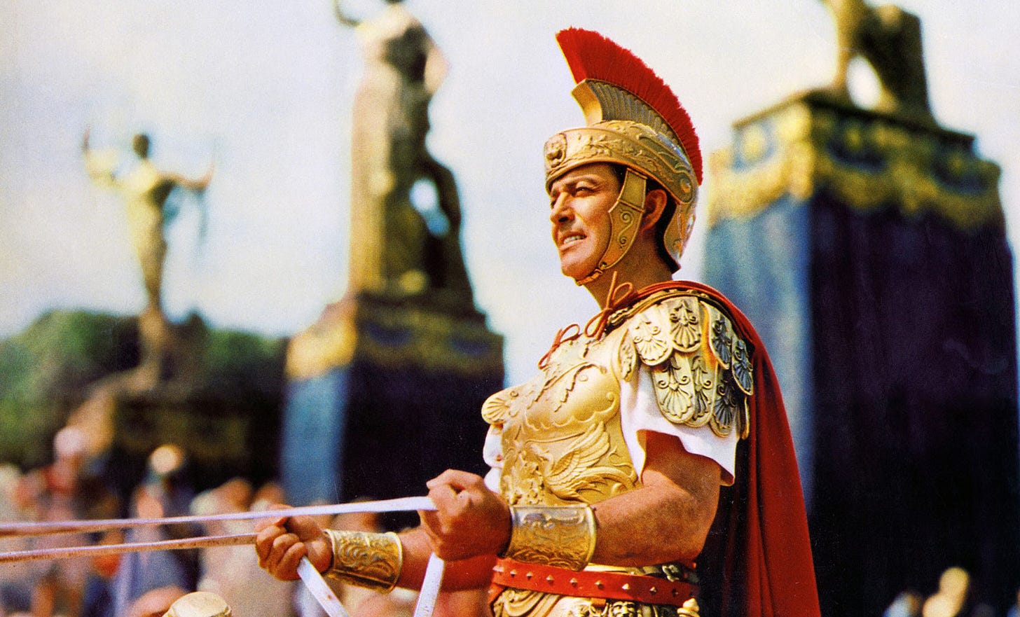 Robert Taylor as Marcus Vinicius in a fabulous, gold armoured uniform with red-crested hat, holds the reins of his chariot in front of grand statues in 1951 biblical epic Quo Vadis