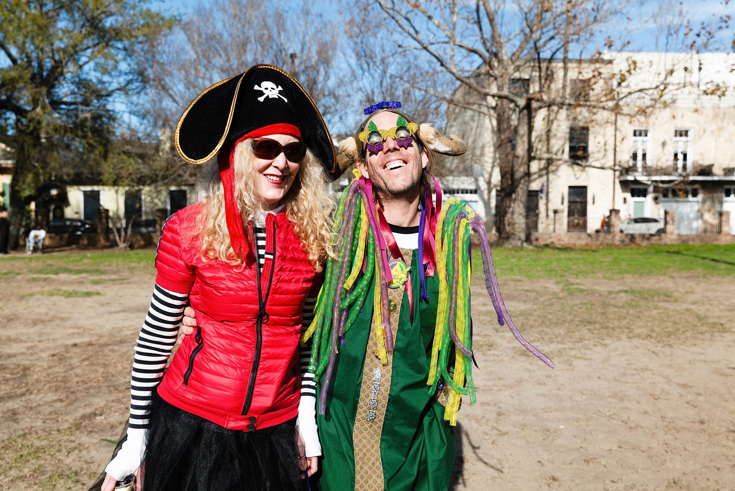 A woman wearing a pirate hat, striped shirt, and red vest, and a man dressed in a zany purple, green, and gold Mardi Gras costume, in a park.