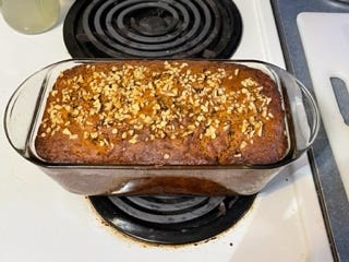 A loaf of breakfast bread in a glass bread pan on an electric stove cooling. It is dark because of the ingredients with a light dusting of walnuts, chopped.