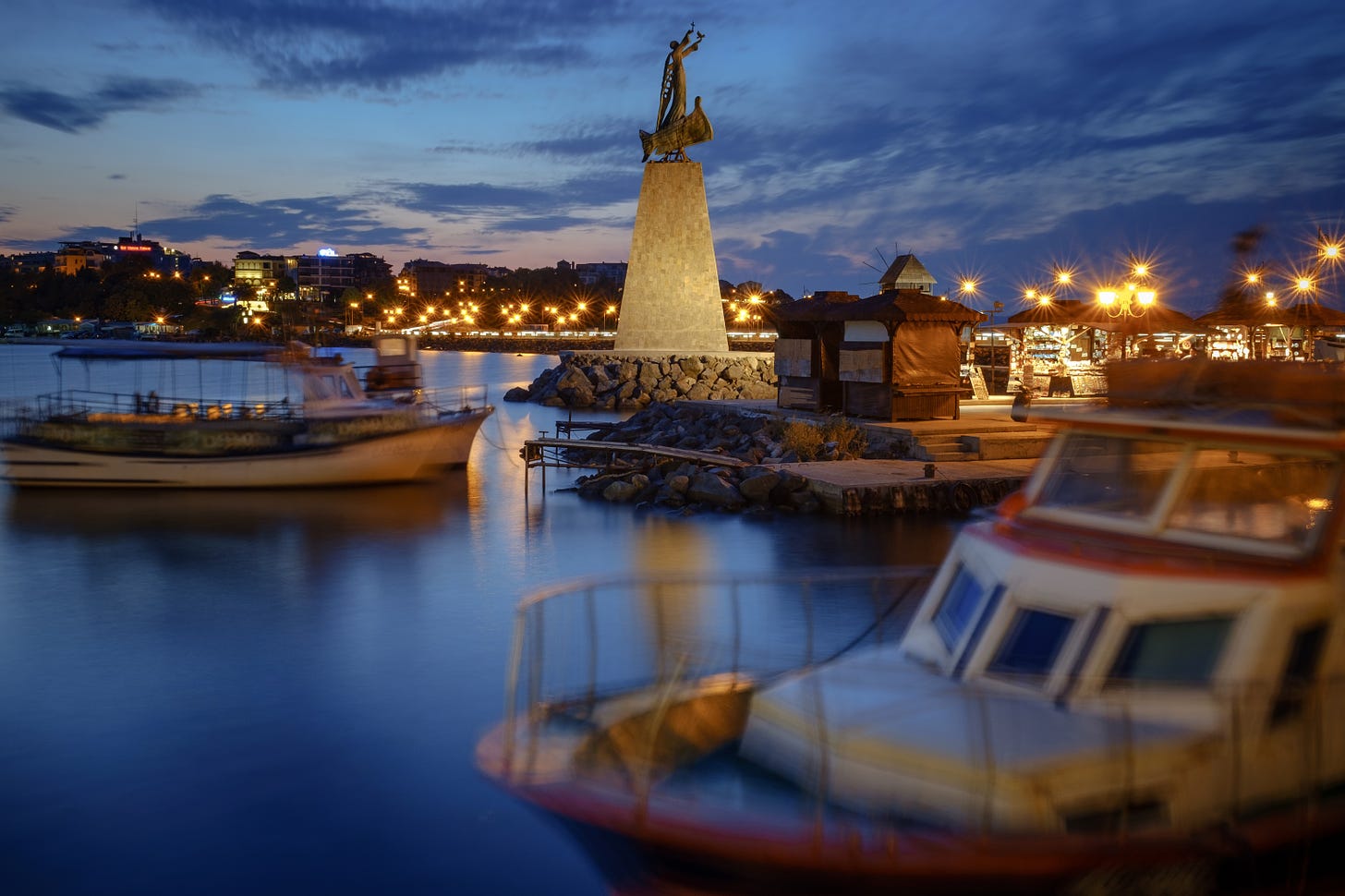 Boats bobbing around in the harbour at dusk. A statue of a person stands on a stone monument.