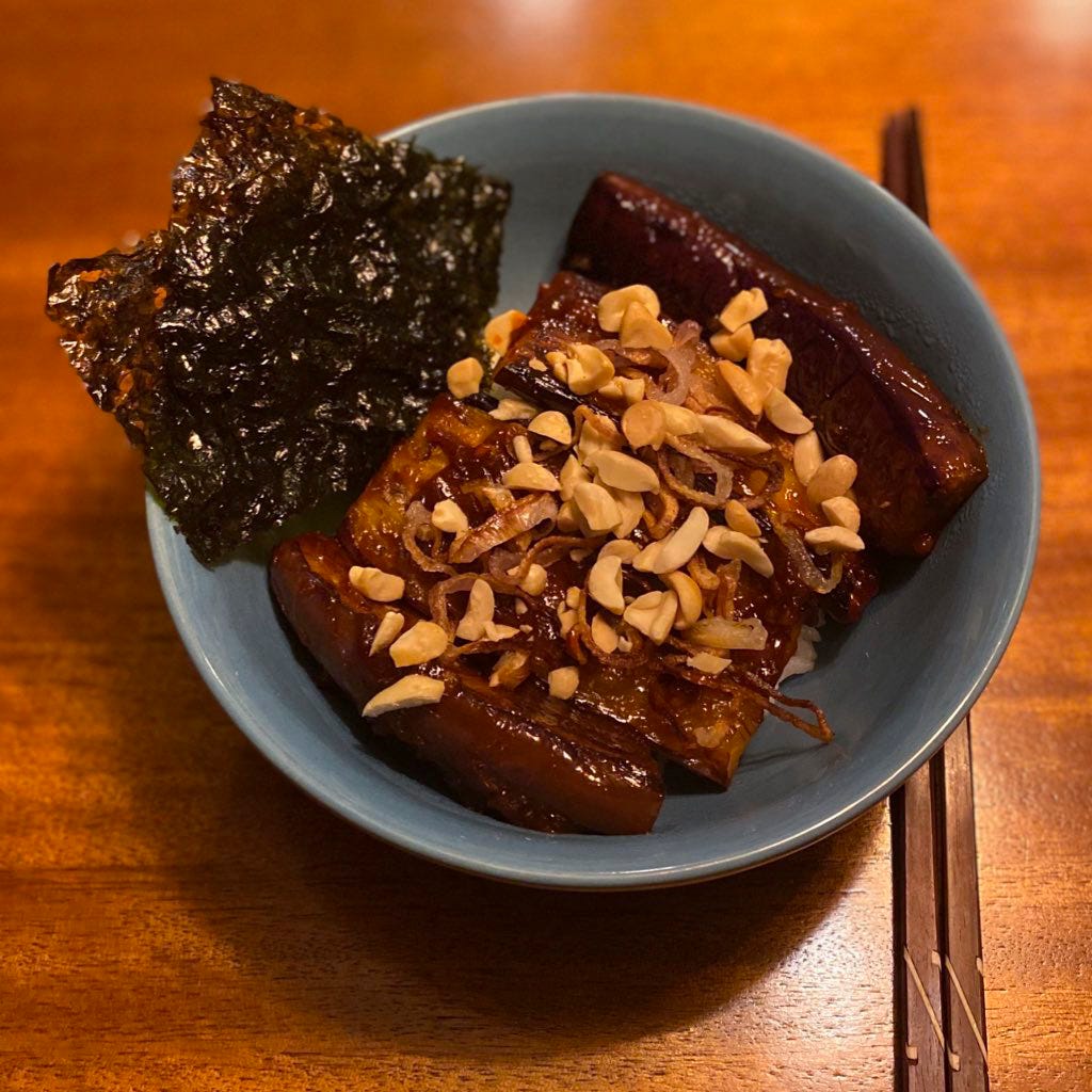 A bowl of rice with four thick slices of eggplant in a deep reddish-brown gochujang glaze. On top are crisp slices of shallot and chopped peanuts, and at the edge of the bowl are two sheets of gim. Chopsticks rest on the table beside it.