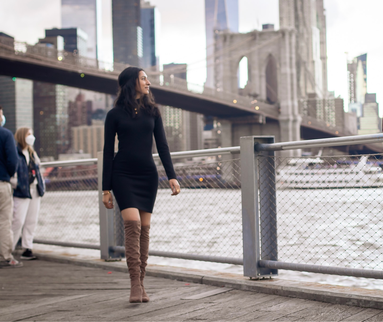 Abi in a black dress and brown boots walking towards the camera in front of a blurry Brooklyn Bridge.
