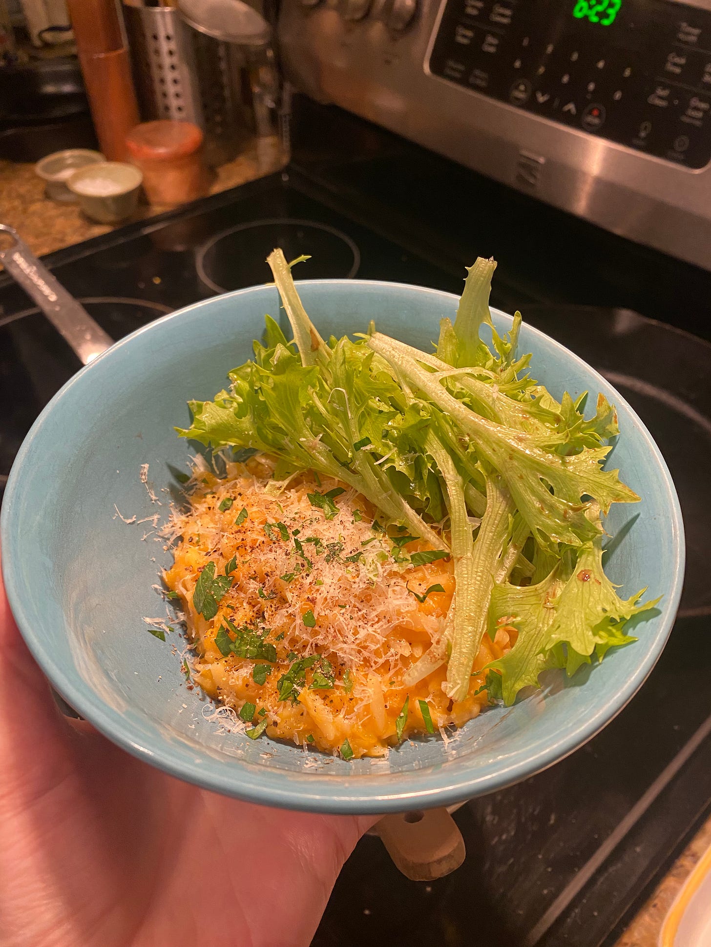 The orzo dish described above, in a blue bowl. It's creamy and orange with sprinkles of parsley and finely grated cheese on top. A small amount of frisée salad covers one half of the bowl. I am holding it in front of the stove.