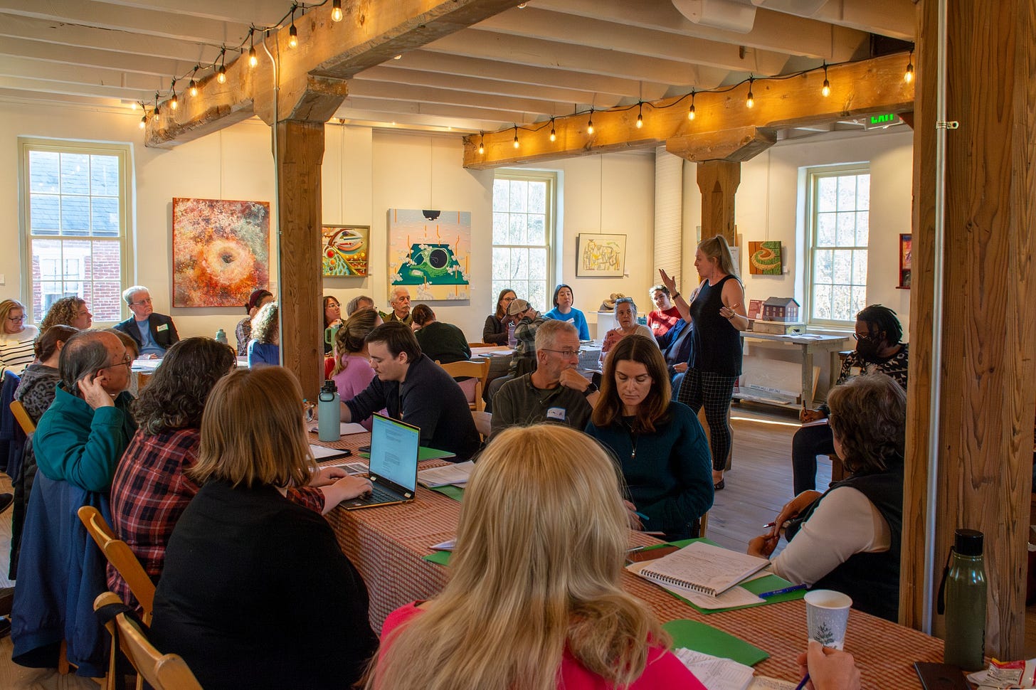 A room of writers sitting in an L-shape. I’m standing toward the left, leading a workshop. A room of writers sitting in an L-shape. I’m standing toward the left, leading a workshop.