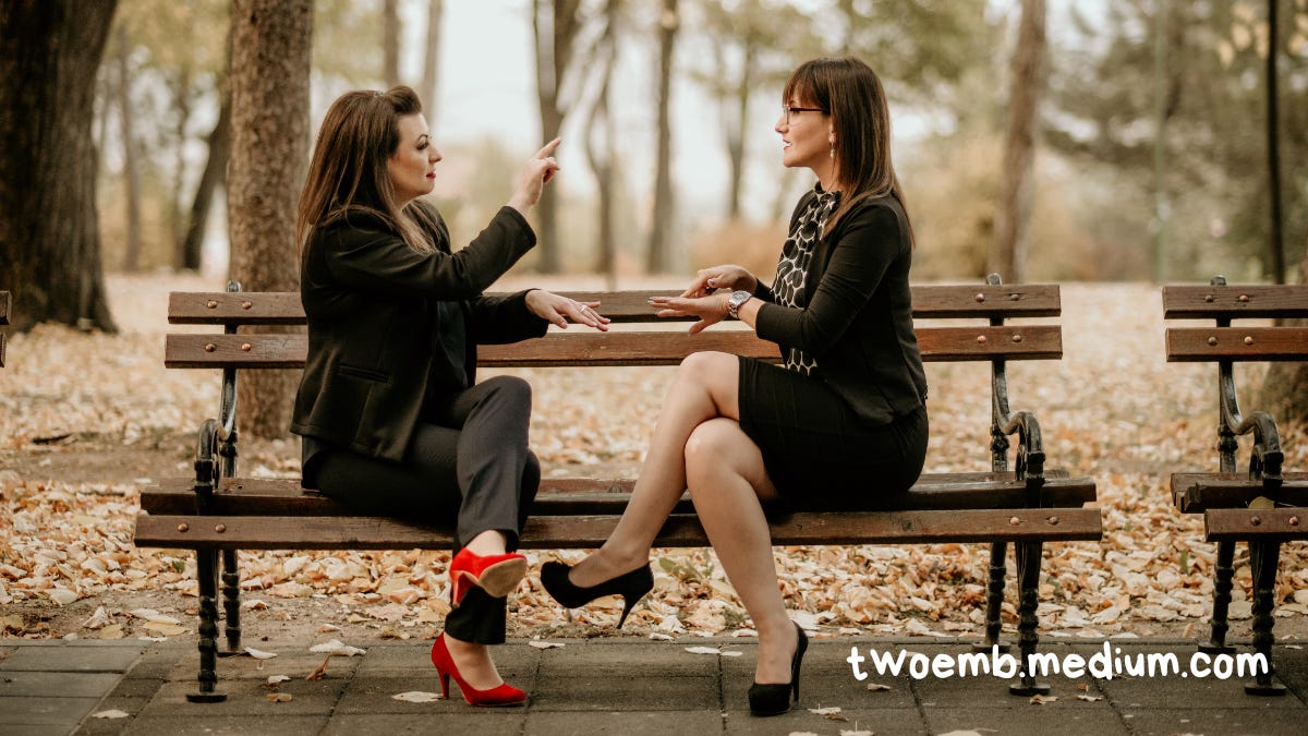 Two women are sitting on a park bench. One woman, wearing red shoes, is fingerspelling. The other woman is signing “slow”, asking her to spell slowly.