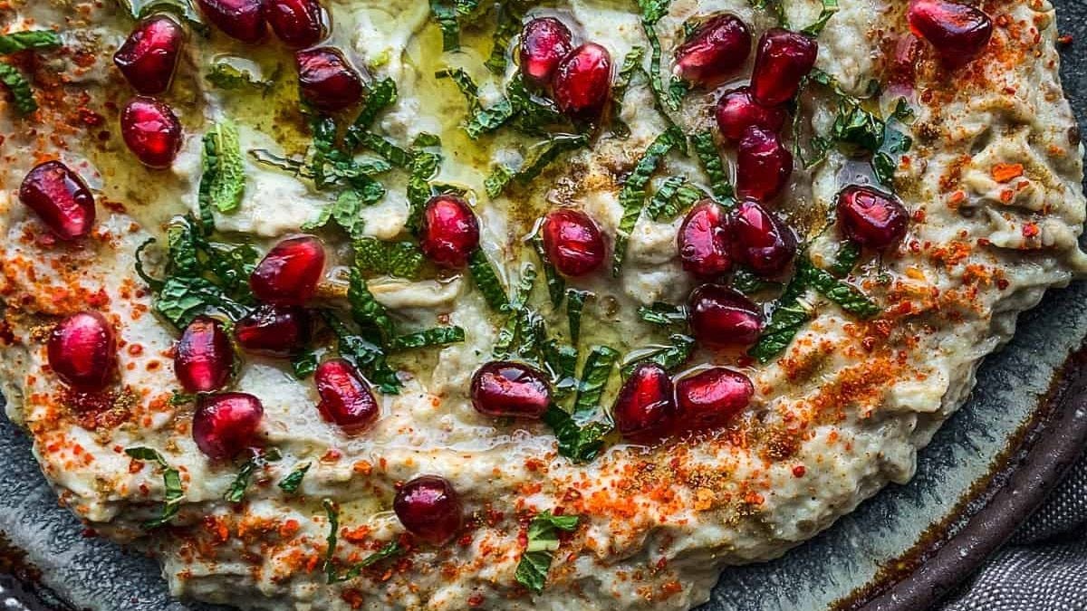 A dish of baba ganoush garnished with pomegranate seeds, chopped herbs, olive oil, and a sprinkle of red spice, served on a rustic plate.