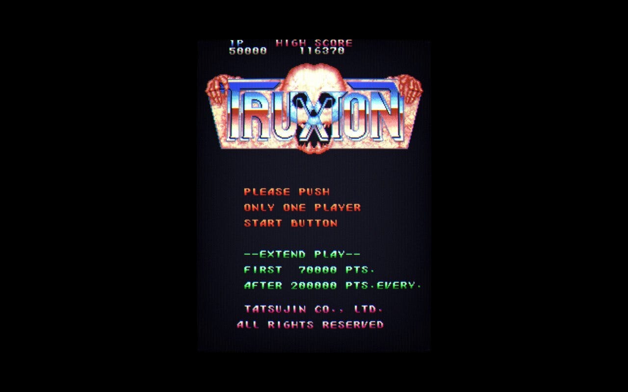 A screenshot of Truxton's title screen, featuring its skull logo with skeleton hands reaching out from behind it over the top. The background is all black, while the skull and logo have a pinkish hue to them, and the word Truxton itself is colored with a blue and brown gradient.