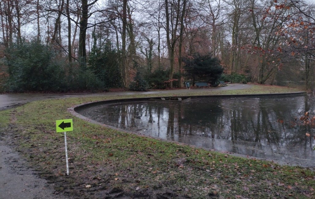 A pond is beside the course, with a muddy grassy verge. The course goes by on tarmacced paths.