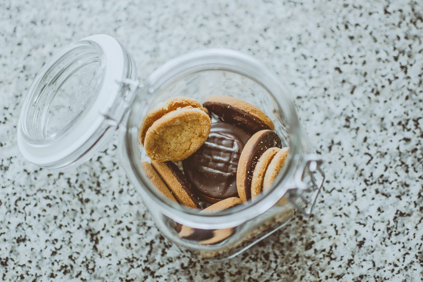 A clear glass cookie jar on a white and black table. The camera is looking down into the jar and inside are yellow cookies with chocolate in the middle.