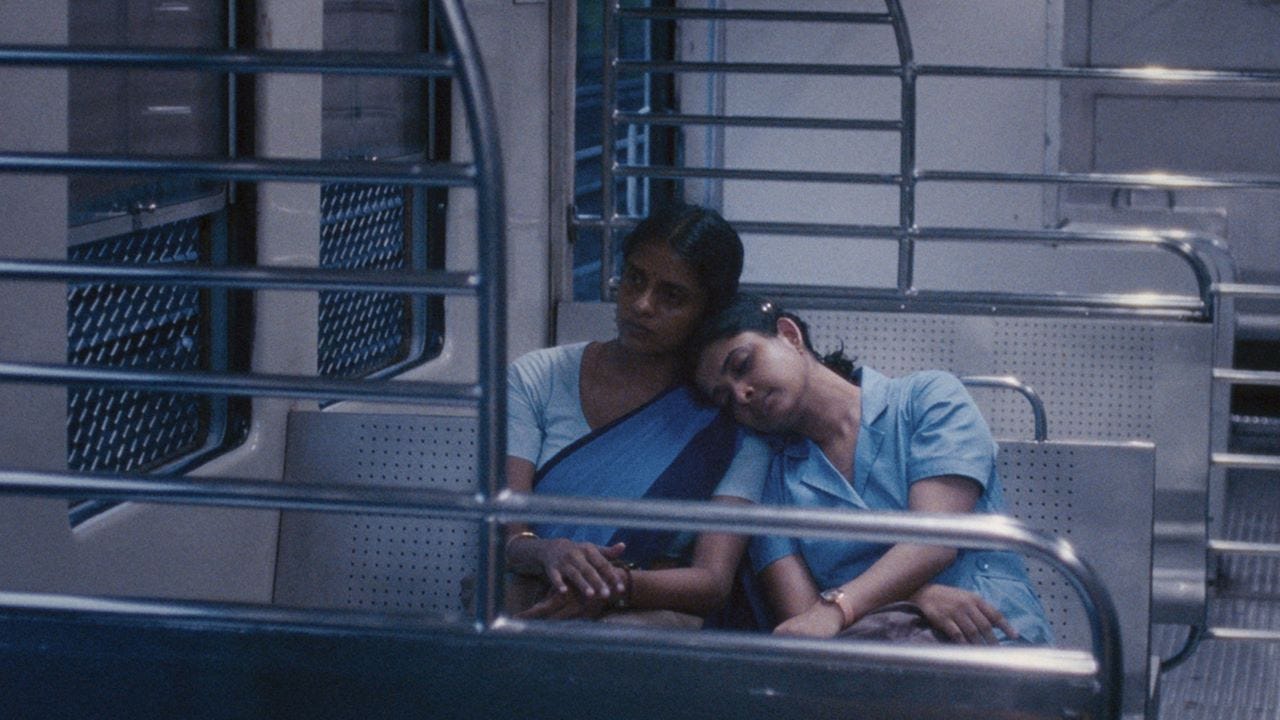 Two women, Prabha (Kani Kusruti) and Anu (Divya Prabha) sitting on an empty bus wearing saris. Anu is resting her head on Prabha's shoulder.