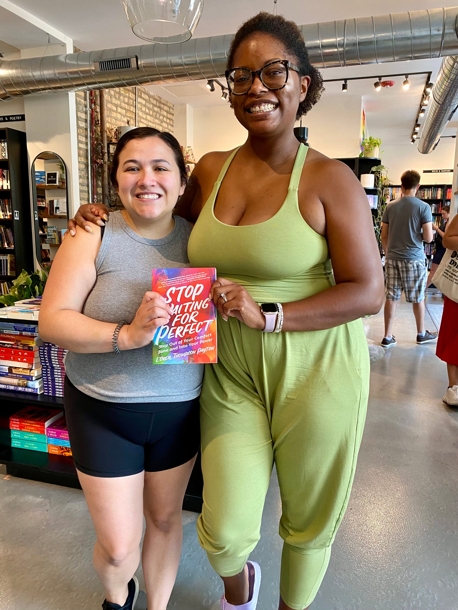 Picture of two women holding a book inside of a bookstore. One is wearing a grey top and black pants and the other is wearing a green jumpsuit
