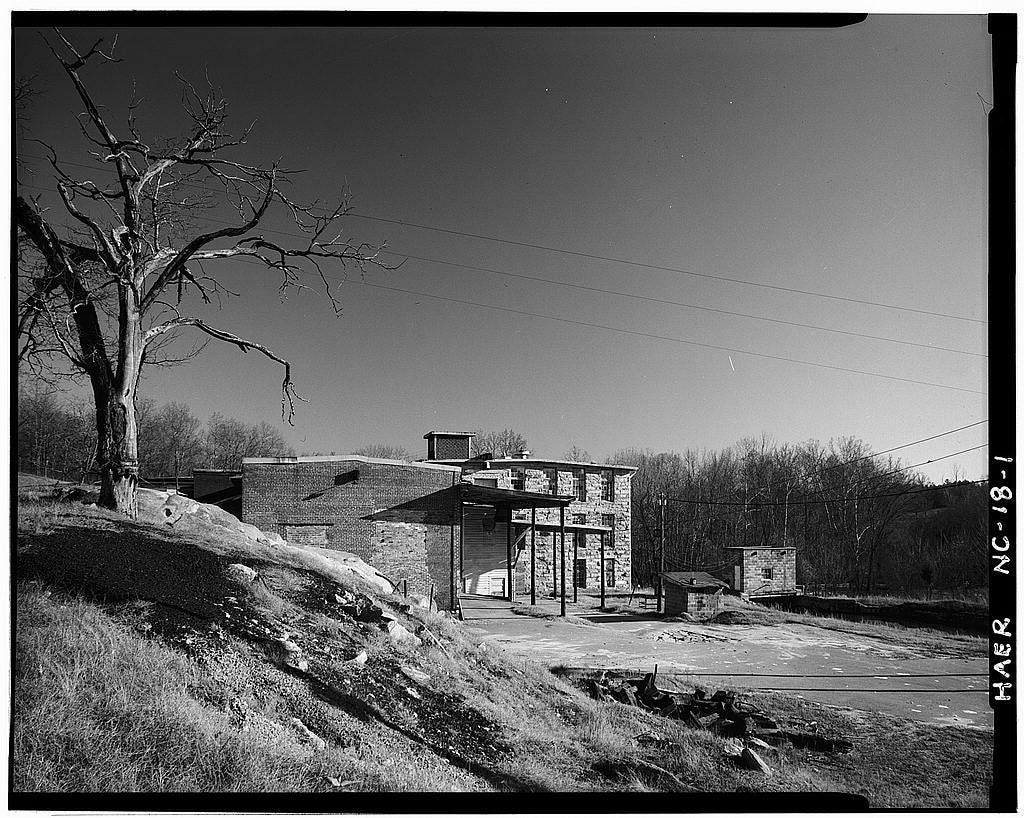 black and white photograph of mill building with river rushing from the dam; a bare tree on a hill overlooking